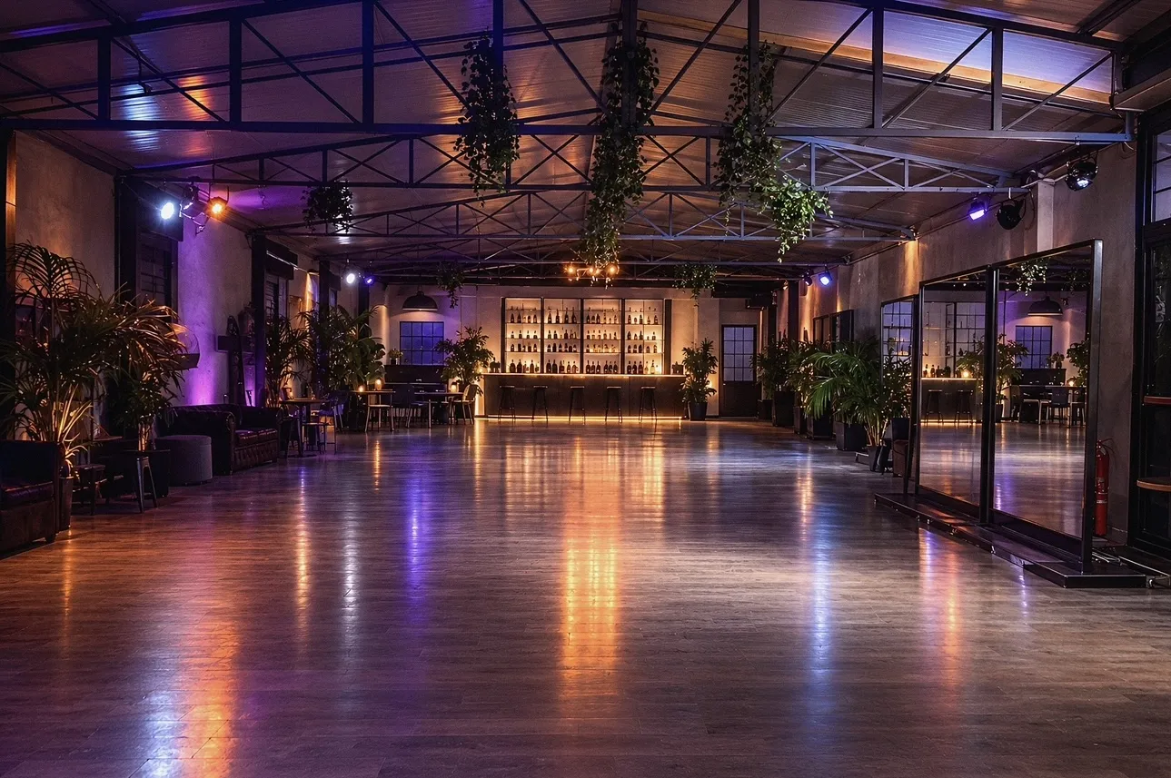 Sala da ballo con travi in ferro, bar illuminato e specchi, luci viola e ambra